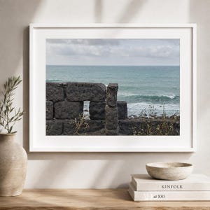 Puede incluir: Una fotografía de paisaje enmarcada que muestra un muro de piedra gris oscuro con una pequeña abertura rectangular, frente a un océano turquesa con olas suaves bajo un cielo nublado. La impresión se exhibe en un marco blanco en la pared.