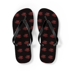 May include: A pair of black flip-flops with a repeating pattern of the letters "AL" in a reddish-pink script. The thong straps are black, and the background is black.
