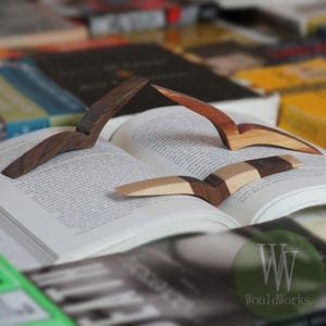May include: Three wooden page holders rest on an open book. The page holders are made of different types of wood, including dark brown, light brown, and a combination of both. The book is surrounded by other books, creating a cozy reading scene.