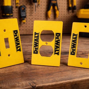 May include: Three yellow electrical outlet covers with the "DEWALT" logo in black. The covers are for a light switch, a standard outlet, and a rectangular opening. The covers are on a wooden surface.