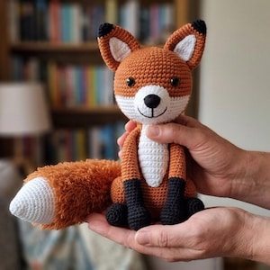 Puede incluir: Muñeco de zorro de ganchillo hecho a mano con detalles en naranja, blanco y negro. El zorro tiene una barriga blanca, una nariz y patas negras y una cola naranja esponjosa. El juguete está en manos de alguien.