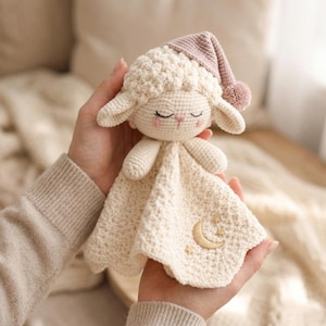 Puede incluir: Una manta de seguridad de cordero de ganchillo hecha a mano en crema con un gorro de dormir rosa. El cordero tiene los ojos cerrados, una nariz rosa y pestañas bordadas. La manta presenta una luna creciente y estrellas bordadas. Un artículo suave y reconfortante para bebés y niños pequeños.