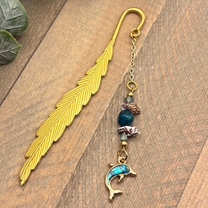 May include: A gold-colored metal bookmark shaped like a feather with a dangling chain attachment. The chain features small blue beads, two miniature seashells, and a gold-toned dolphin charm with an iridescent blue inlay on its body.
