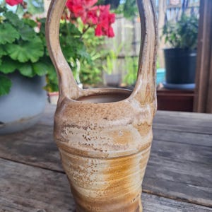 May include: A tall, cylindrical ceramic vessel with a high, arched handle extending from the top. The pottery features a mottled, earthy tan and brown glaze with visible horizontal striations and a textured, speckled finish throughout the surface.