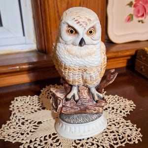 May include: A ceramic owl figurine perched on a brown tree branch base. The owl features white and tan feathers, a black beak, and bright yellow eyes. It stands on a circular white pedestal base with a textured, scalloped edge design.