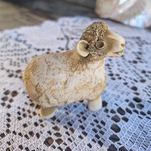 Pode incluir: Uma pequena estatueta de ovelha esculpida à mão em material texturizado de cor clara. A ovelha apresenta uma cabeça detalhada com textura encaracolada e um corpo liso e manchado em tons de castanho e creme, sobre quatro pernas curtas.