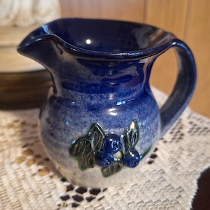 Pode incluir: Um pequeno jarro de cerâmica com um esmalte azul-escuro brilhante no topo, que transita para uma base azul-clara e branca salpicada. A frente apresenta um relevo tridimensional de mirtilos azul-escuros com folhas verdes.