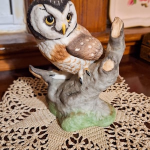 May include: A ceramic owl figurine perched on a textured tree branch. The owl features white, brown, and black plumage with bright yellow eyes and a yellow beak. The base of the branch is painted with a light green wash, and the owl has black talons.