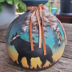 May include: A round matte black vase featuring a painted silhouette of a wolf standing in a forest against a gradient background of yellow, teal, and dark blue. The neck is adorned with a braided tan leather cord and several brown feathers.