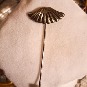 Può includere: Una spilla da bavero color argento con una parte superiore a forma di ventaglio e dettagli a coste verticali. La lunga e sottile spilla in metallo termina con un piccolo tappo arrotondato alla base, per una finitura sicura dell'accessorio.