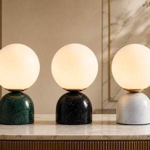 Puede incluir: Tres lámparas de mesa modernas con pantallas esféricas de vidrio blanco. Las lámparas tienen una base dorada y se asientan sobre una base de mármol verde, negro y blanco. Las lámparas están dispuestas sobre una superficie de mármol.