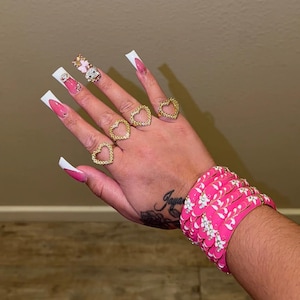 Puede incluir: Una mano con anillos dorados en forma de corazón y una pulsera rosa con estampado floral. Las uñas largas tienen un diseño blanco y rosa, y una uña tiene un adorno.