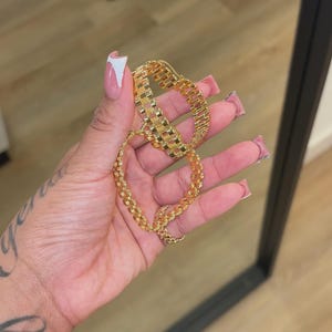 Puede incluir: Un conjunto de pulsera y collar de color dorado. Las joyas presentan un diseño geométrico texturizado. La pulsera está abrochada y el collar es una cadena. Las joyas se sostienen en una mano con manicura.