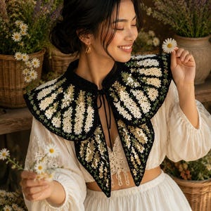 Peut inclure: Un col en crochet noir orné d'un motif décoratif de fleurs blanches et vertes avec de subtiles paillettes. Le col présente une encolure arrondie et une fermeture à nouer sur le devant avec deux longs cordons noirs qui pendent.