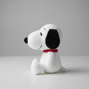 May include: A handmade white crochet dog with black ears and a black nose. The dog is wearing a red collar and is sitting down. The dog is a character from the Peanuts comic strip.