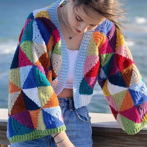 May include: A colorful, patchwork crochet cardigan with a light blue trim. The cardigan features a variety of geometric shapes in shades of red, blue, green, yellow, and orange. The sleeves are long and the cardigan is open in the front.