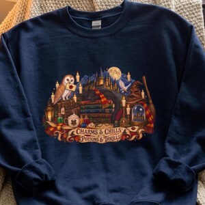 May include: A navy blue crewneck sweatshirt featuring a detailed graphic print of a stack of books, a barn owl, a black cat, a broomstick, and a castle silhouette. The text "CHARMS & CHILLS POTIONS & THRILLS" is printed on a banner at the bottom.
