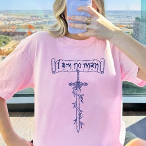 May include: A light pink t-shirt with the text "I am no man" above a sword design. The sword is wrapped with vines. The word "BLOSSOM" is in a black box at the bottom of the image.