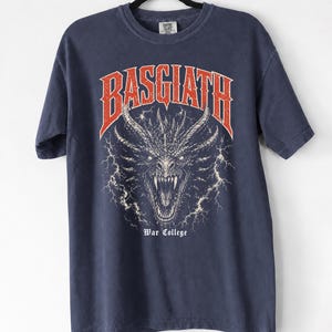 Puede incluir: Camiseta azul marino con un dise&ntilde;o de drag&oacute;n y la palabra "BASGIATH" en rojo encima. El texto "War College" est&aacute; debajo del drag&oacute;n. La camiseta est&aacute; colgada en una percha negra.