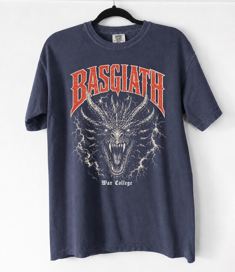 Puede incluir: Camiseta azul marino con un dise&ntilde;o de drag&oacute;n y la palabra "BASGIATH" en rojo encima. El texto "War College" est&aacute; debajo del drag&oacute;n. La camiseta est&aacute; colgada en una percha negra.