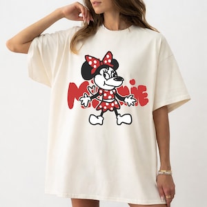 May include: Cream-colored t-shirt featuring a cartoon Minnie Mouse design. Minnie is depicted in a red polka-dot dress and bow, with the name "Minnie" in red script. The shirt has short sleeves and a relaxed fit.