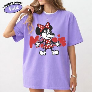 May include: A violet Comfort Colors t-shirt featuring a Minnie Mouse graphic. The cartoon character is depicted in a red and white polka-dot dress and bow, with the name "Minnie" in red letters. The shirt has short sleeves.