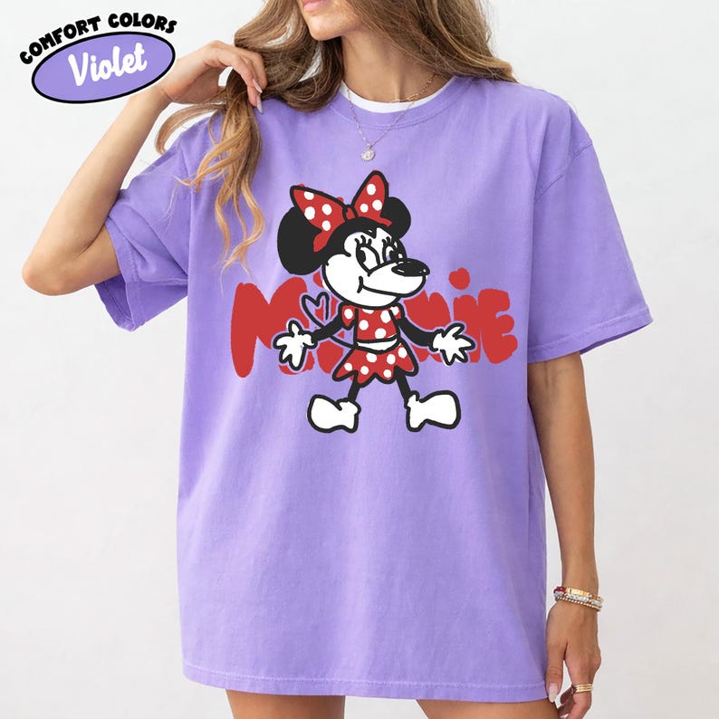 May include: A violet Comfort Colors t-shirt featuring a Minnie Mouse graphic. The cartoon character is depicted in a red and white polka-dot dress and bow, with the name "Minnie" in red letters. The shirt has short sleeves.