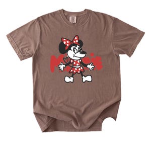 May include: Brown t-shirt featuring a cartoon Minnie Mouse graphic. Minnie is depicted in a red polka-dot dress and bow, with the name "Minnie" in red letters. The shirt has a crew neck and short sleeves.
