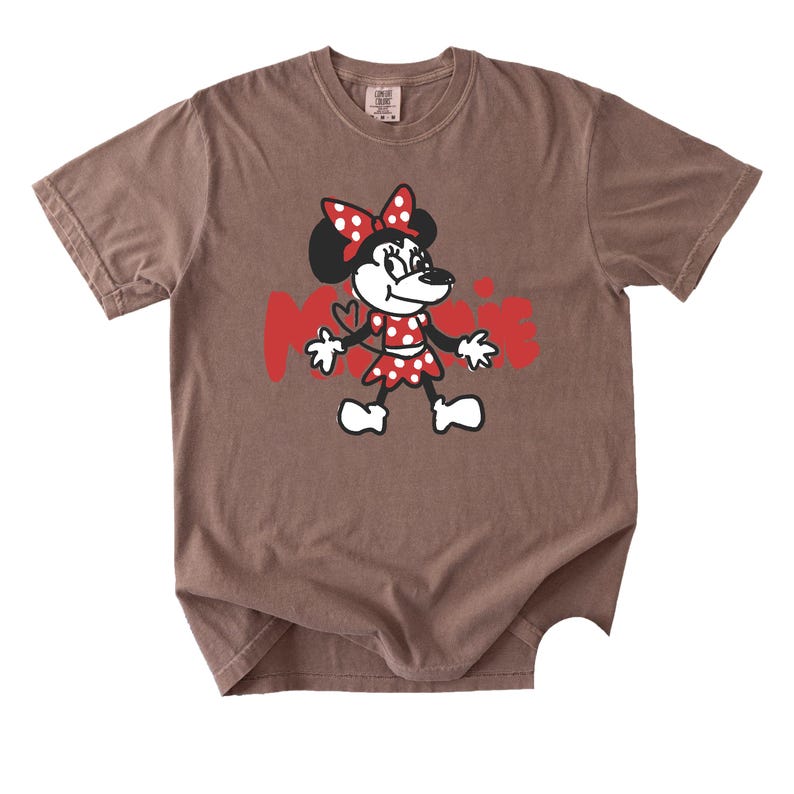 May include: Brown t-shirt featuring a cartoon Minnie Mouse graphic. Minnie is depicted in a red polka-dot dress and bow, with the name "Minnie" in red letters. The shirt has a crew neck and short sleeves.