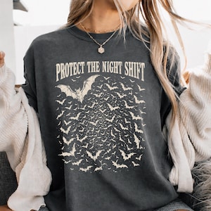 May include: Dark grey t-shirt featuring the text "PROTECT THE NIGHT SHIFT" and a bat graphic with a swarm of bats. The design has a vintage, distressed appearance.