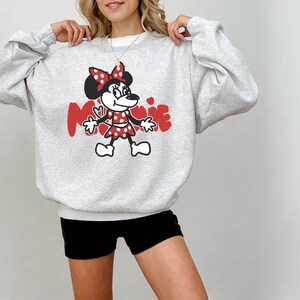May include: A light gray sweatshirt featuring a Minnie Mouse graphic. The graphic includes a red bow, dress, and the name "Minnie" in red. The sweatshirt has long sleeves and a crew neck.