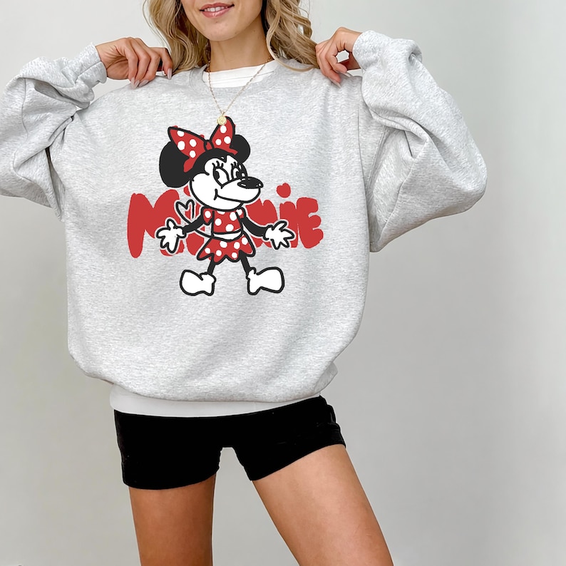 May include: A light gray sweatshirt featuring a Minnie Mouse graphic. The graphic includes a red bow, dress, and the name "Minnie" in red. The sweatshirt has long sleeves and a crew neck.