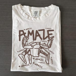 Könnte beinhalten: Cremefarbenes T-Shirt mit brauner Grafik einer muskulösen Figur und dem Wort "AMAZE" in fetten Buchstaben. Das Wort "amaze" wiederholt sich um die Figur. Das Shirt liegt gefaltet auf einer dunklen Holzoberfläche.