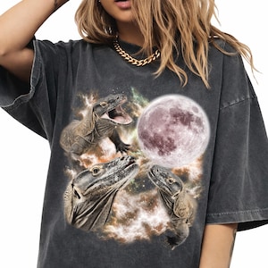 May include: A charcoal gray short-sleeve t-shirt featuring a graphic print of three Komodo dragons surrounding a large, textured full moon against a backdrop of swirling, colorful cosmic nebula clouds and scattered stars.
