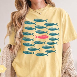May include: A pale yellow short-sleeve t-shirt featuring a graphic print of a school of fish. The design consists of numerous teal-colored fish silhouettes arranged in rows, with a single bright pink fish positioned in the center.