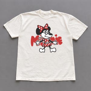 May include: Off-white t-shirt featuring a graphic of Minnie Mouse in a red polka-dot dress and bow. The word "Minnie" is printed in red, with a heart accent. The shirt has short sleeves and a crew neck.