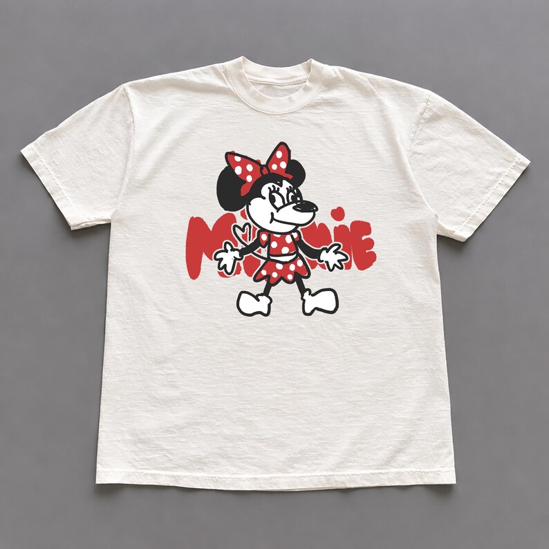 May include: Off-white t-shirt featuring a graphic of Minnie Mouse in a red polka-dot dress and bow. The word "Minnie" is printed in red, with a heart accent. The shirt has short sleeves and a crew neck.