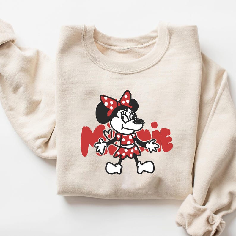 May include: A beige sweatshirt featuring a cartoon illustration of Minnie Mouse in a red polka-dot dress and bow. The word "Minnie" is printed in red, stylized lettering. The sweatshirt is folded.