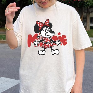 May include: Cream-colored t-shirt featuring a cartoon Minnie Mouse design. Minnie is depicted in a red polka-dot dress and bow, with the word "MINNIE" in red block letters. The design is centered on the shirt.