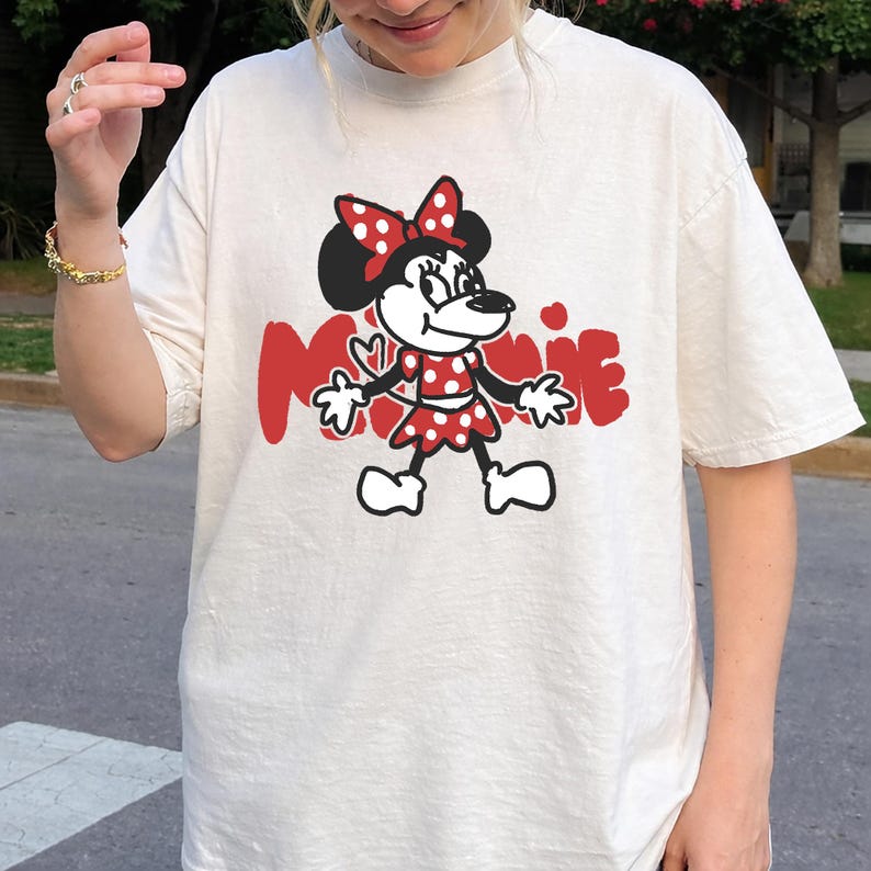 May include: Cream-colored t-shirt featuring a cartoon Minnie Mouse design. Minnie is depicted in a red polka-dot dress and bow, with the word "MINNIE" in red block letters. The design is centered on the shirt.