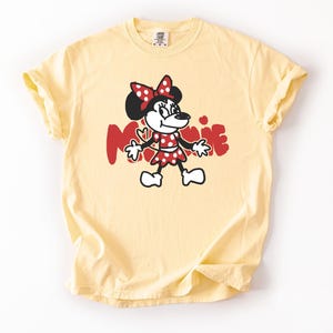 May include: A light yellow t-shirt featuring a cartoon illustration of Minnie Mouse. Minnie is depicted in a red polka-dot dress and bow, with the name "Minnie" in red letters. The shirt has short sleeves and a crew neck.