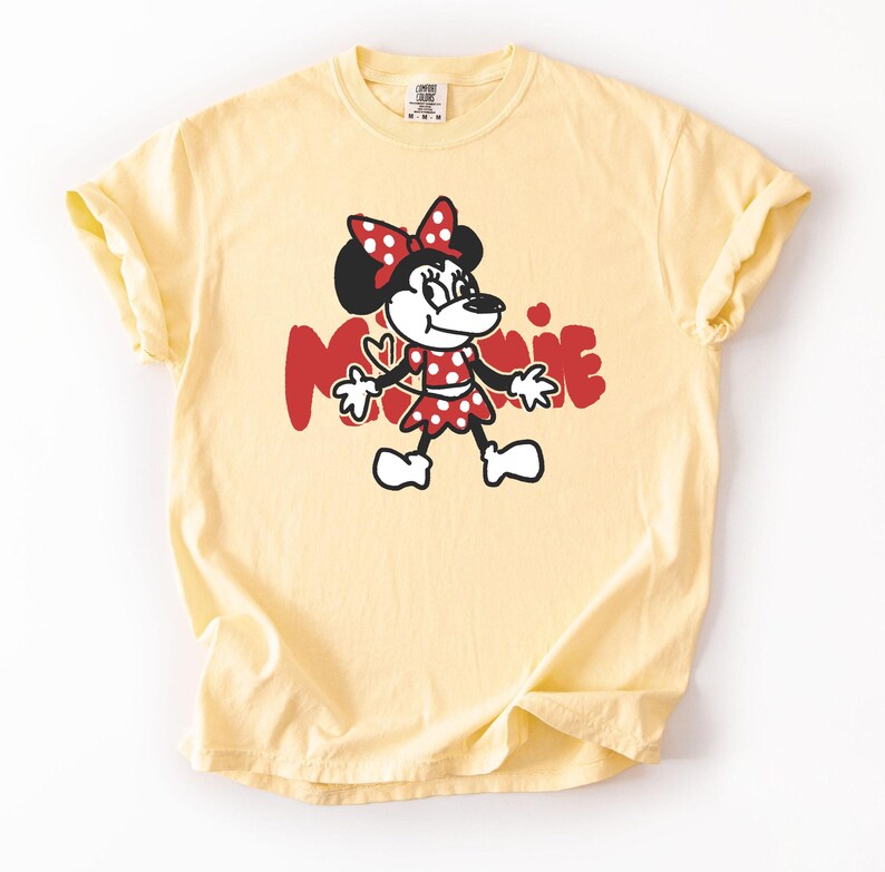 May include: A light yellow t-shirt featuring a cartoon illustration of Minnie Mouse. Minnie is depicted in a red polka-dot dress and bow, with the name "Minnie" in red letters. The shirt has short sleeves and a crew neck.