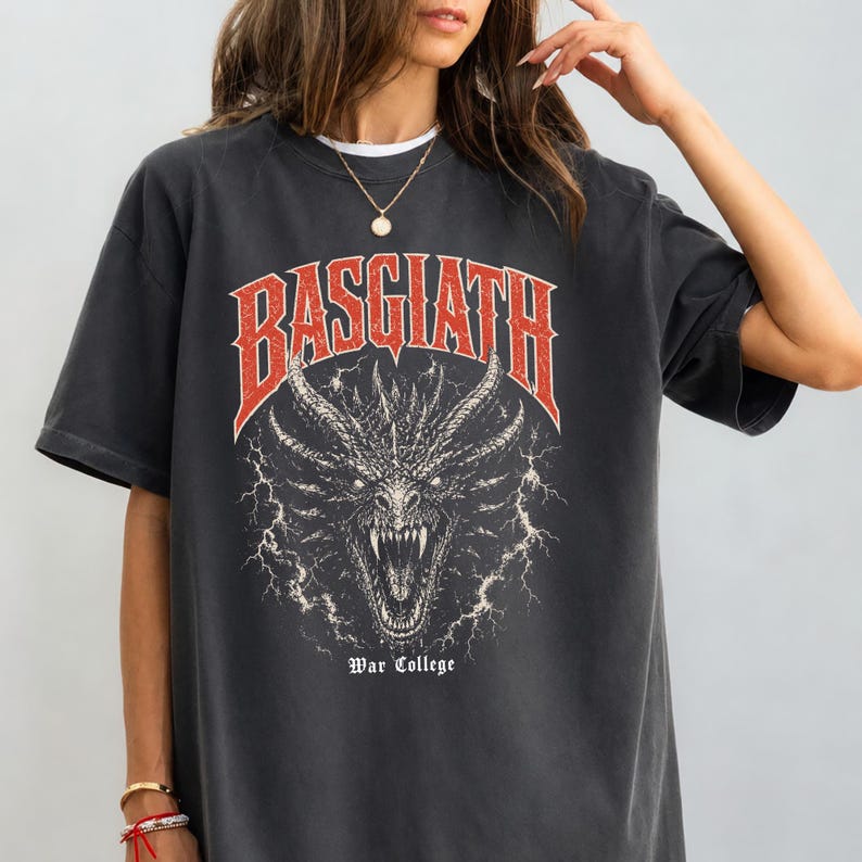 Puede incluir: Camiseta gris oscuro con un dise&ntilde;o de drag&oacute;n y la palabra "BASGIATH" en rojo arriba. El texto "War College" est&aacute; debajo. El drag&oacute;n tiene dientes afilados y est&aacute; rodeado de rayos. Una camiseta gr&aacute;fica informal.