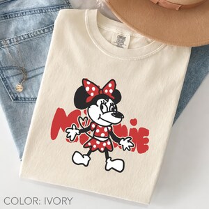 May include: Ivory-colored t-shirt featuring a cartoon Minnie Mouse design in red and black. The graphic includes Minnie's name in red script. The shirt is paired with blue jeans and a tan hat.