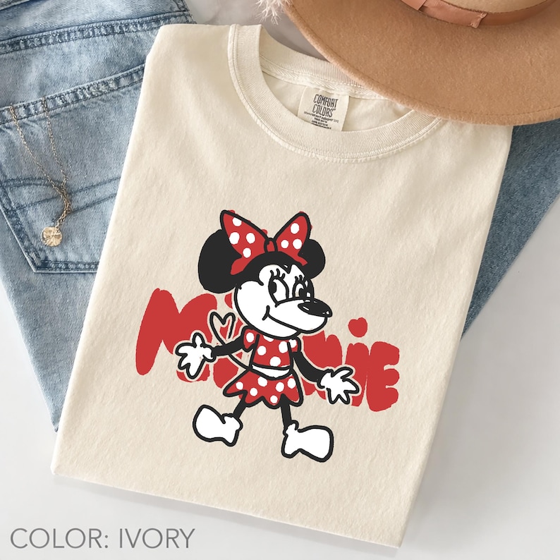 May include: Ivory-colored t-shirt featuring a cartoon Minnie Mouse design in red and black. The graphic includes Minnie's name in red script. The shirt is paired with blue jeans and a tan hat.