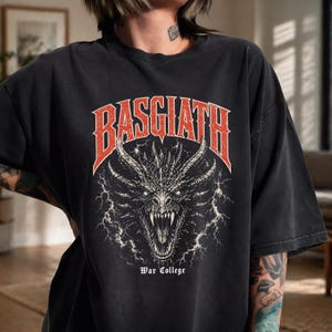 Puede incluir: Camiseta negra con un dise&ntilde;o de drag&oacute;n detallado y la palabra "BASGIATH" en rojo. El texto "War College" est&aacute; debajo. El dise&ntilde;o tiene un aspecto vintage y desgastado, con una ilustraci&oacute;n de drag&oacute;n.