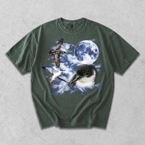 May include: A dark olive green short-sleeve t-shirt featuring a graphic print of a seabird in flight, a close-up of a seabird head, and a full moon set against a backdrop of blue and white clouds and a starry night sky.
