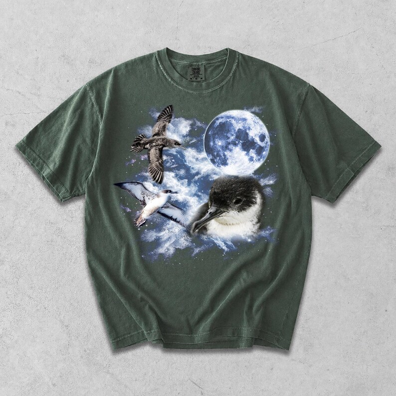 May include: A dark olive green short-sleeve t-shirt featuring a graphic print of a seabird in flight, a close-up of a seabird head, and a full moon set against a backdrop of blue and white clouds and a starry night sky.