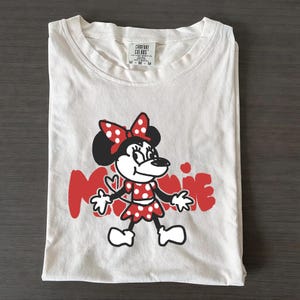 May include: White t-shirt featuring a red and black Minnie Mouse graphic. The design includes Minnie Mouse in a red polka dot dress and bow, with the name "Minnie" in red. The shirt is folded and has a tag at the top.