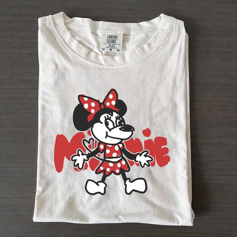 May include: White t-shirt featuring a red and black Minnie Mouse graphic. The design includes Minnie Mouse in a red polka dot dress and bow, with the name "Minnie" in red. The shirt is folded and has a tag at the top.